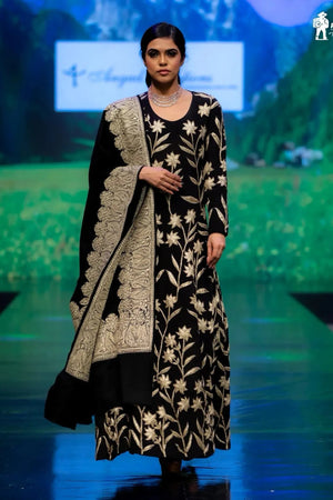 Model wearing a black and gold embroidered outfit on a runway with a scenic backdrop.
