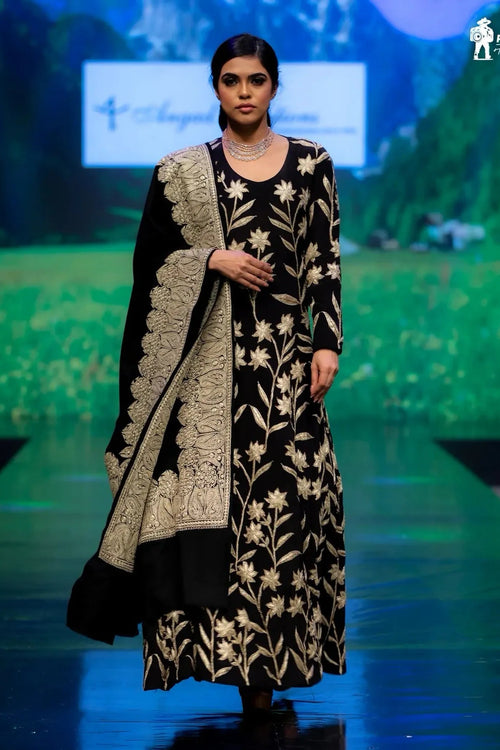 Model wearing a black and gold embroidered outfit on a runway with a scenic backdrop.