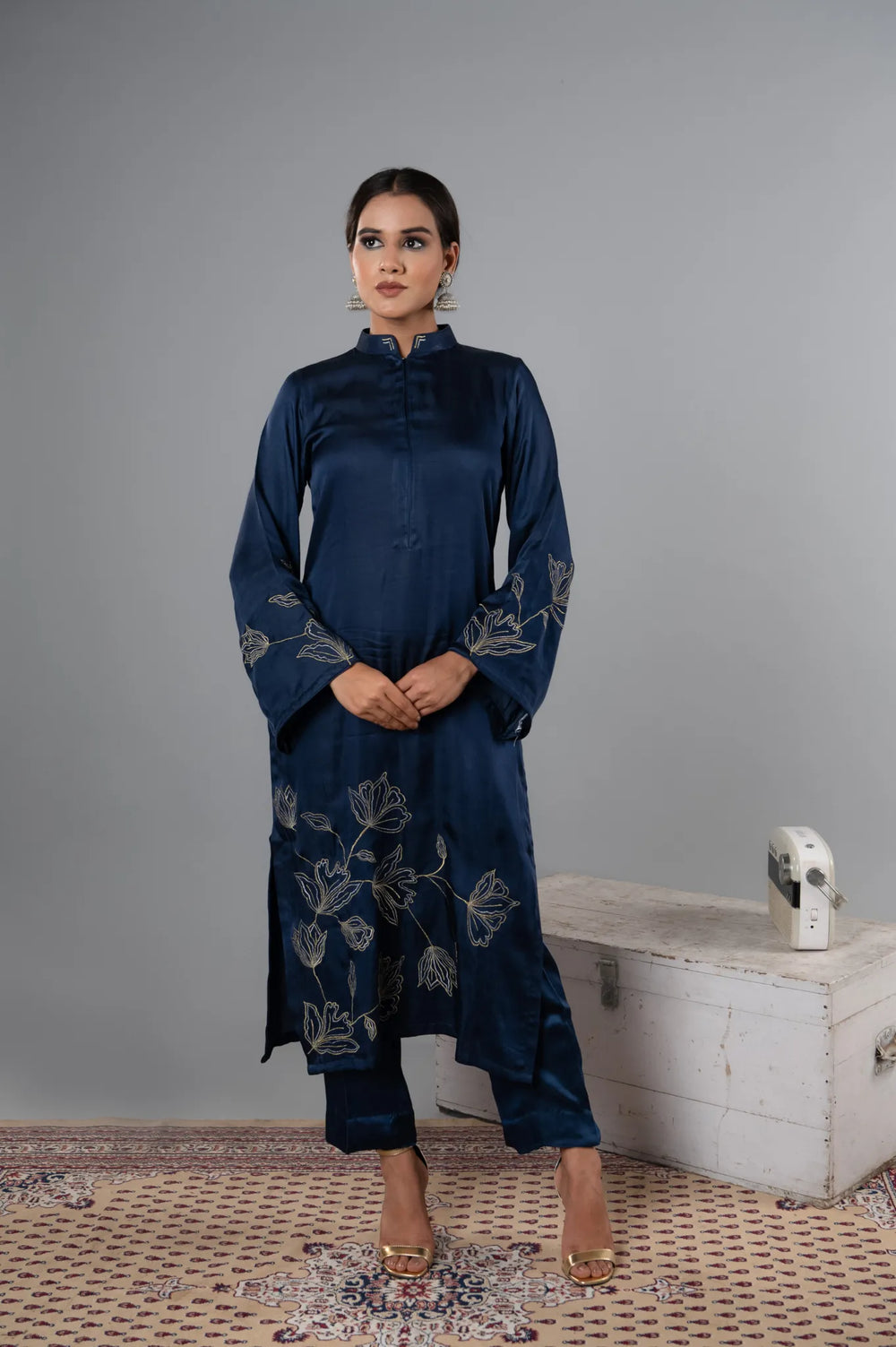 Woman wearing a dark blue embroidered outfit standing against a gray wall.