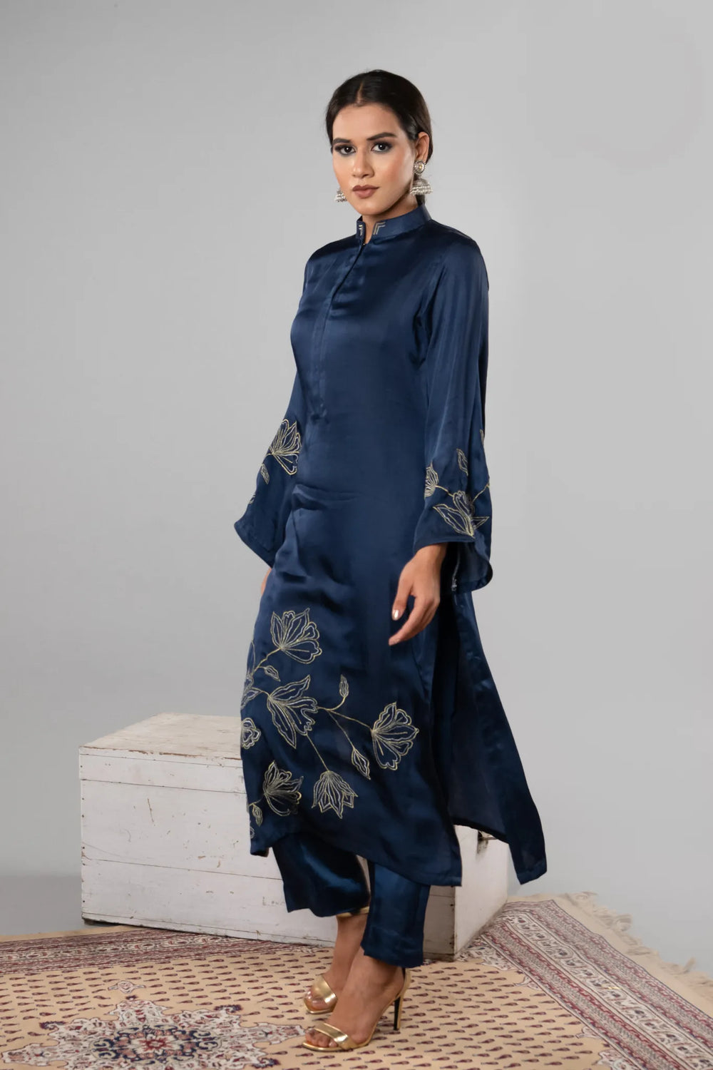 Woman wearing a dark blue embroidered outfit standing against a gray wall.