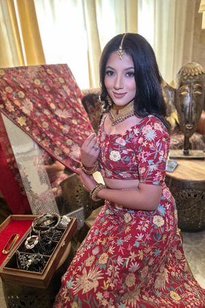 Woman in a red floral traditional outfit posing indoors with decorative items.