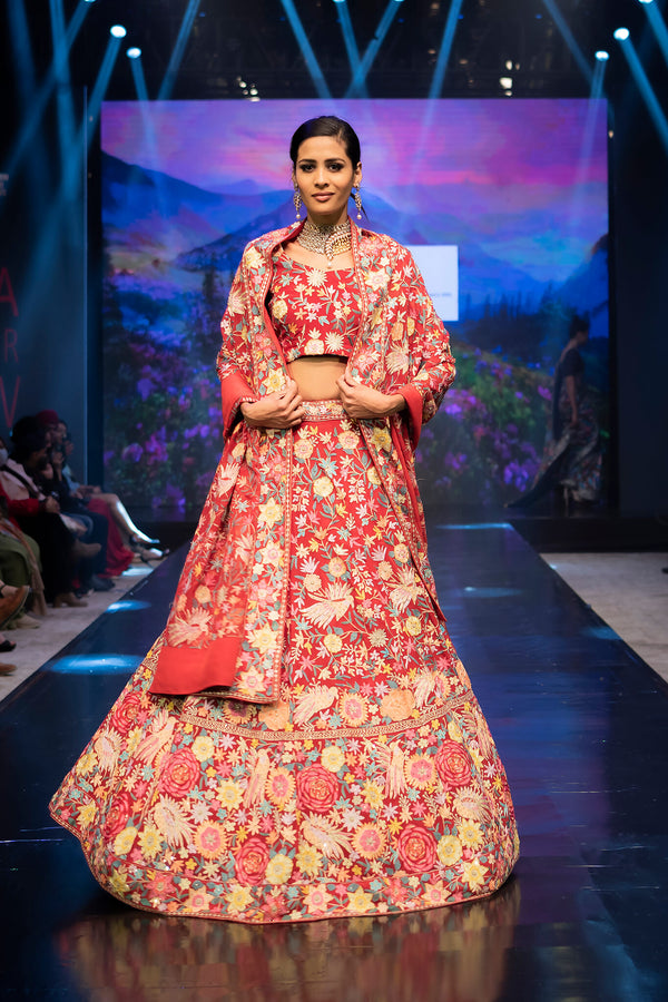 Model wearing a red floral outfit on a fashion show runway with a scenic backdrop.