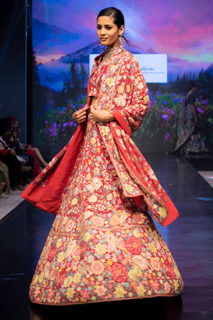 Model wearing a floral and red traditional outfit on a runway with a scenic backdrop