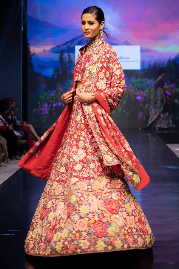 Model wearing a floral and red traditional outfit on a runway with a scenic backdrop