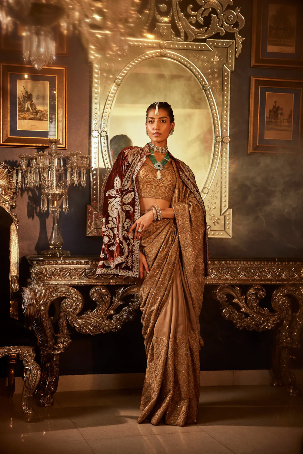 A woman draped in a brown saree with golden and silver sequence work, standing in a luxurious interior setting.
