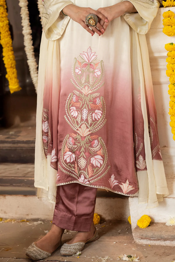 #OmbreCream&Mauve Traditional outfit with intricate embroidery, held by a person, against a decorated background.