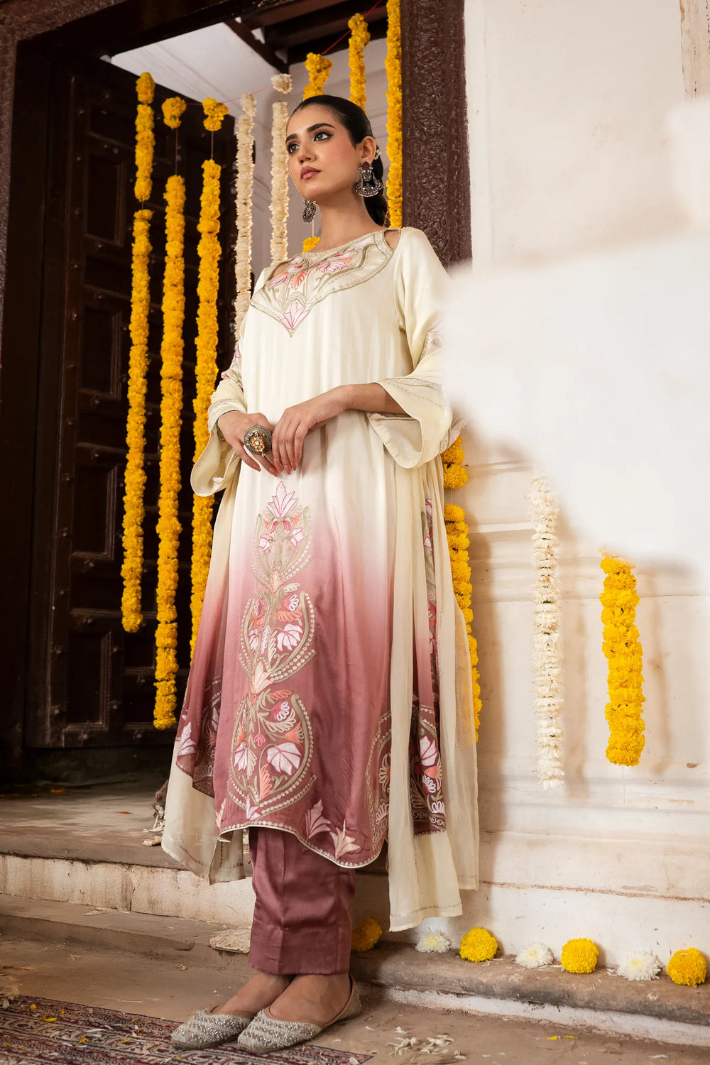 #OmbreCream&Mauve Woman in traditional outfit standing in front of a decorated entrance with yellow flowers.