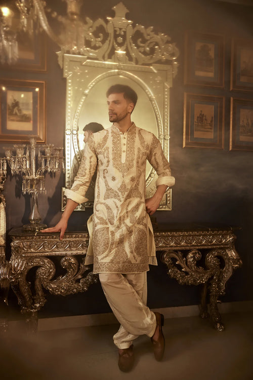 A man wearing a white and golden embroidered kurta pyjama standing in a room with classic decor.