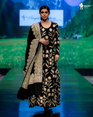 Model wearing a black and gold embroidered outfit on a runway with a scenic backdrop.