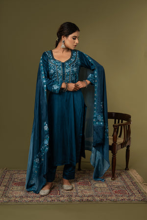 #TealBlue Woman wearing a blue embroidered traditional outfit standing against a plain background Woman in a blue traditional outfit with a cape standing on a patterned rug against a green wall.