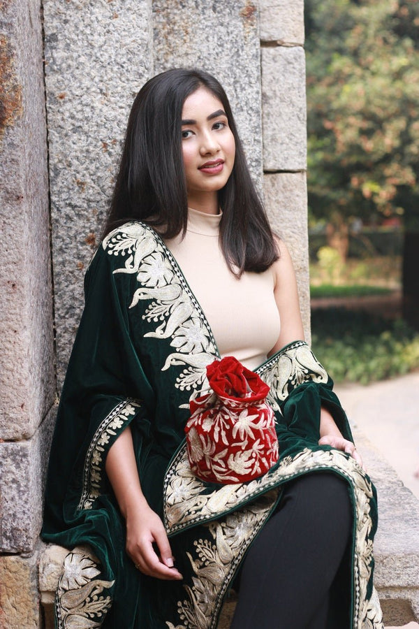 #Red Woman holding a red potli bag in a green and gold shawl against a stone wall.