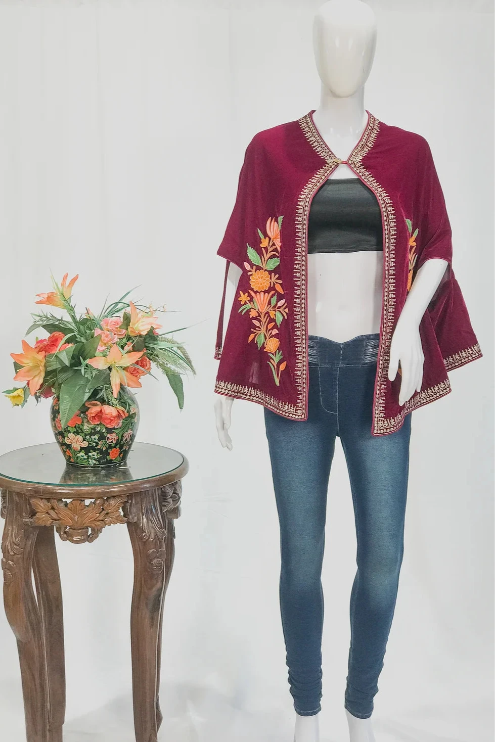 Mannequin wearing a burgundy embroidered top with floral patterns next to a small table with flowers on a white background