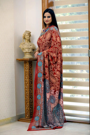 Woman wearing a patterned saree standing indoors with a decorative bust and window blinds in the background.