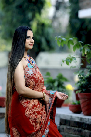 Woman in a red saree with floral patterns standing outdoors with greenery in the background