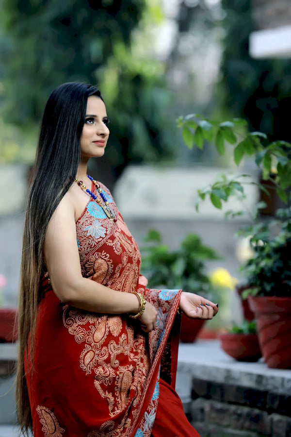 Woman in a red saree with floral patterns standing outdoors with greenery in the background